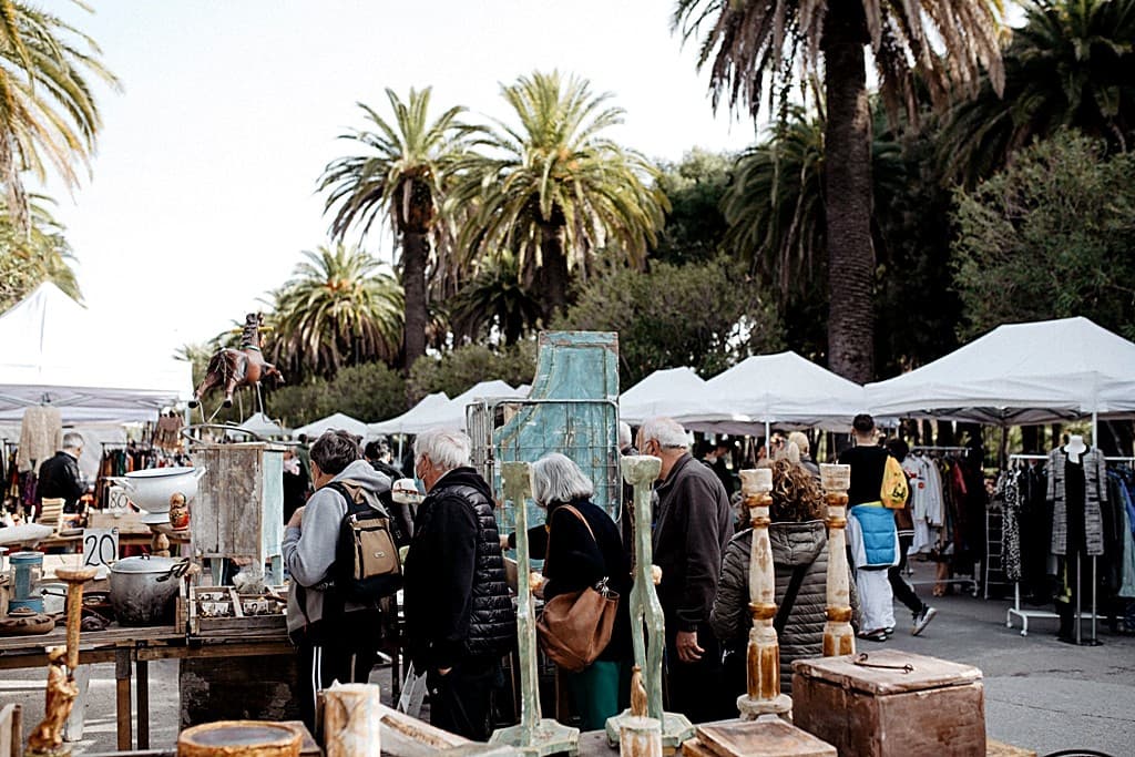 Stand di antiquariato all'edizione di Pasqua di L'Antico e le Palme · candelabri, mobili dipinti, oggetti d'epoca sotto i gazebo bianchi · palme della Riviera sullo sfondo, visitatori che esaminano i pezzi