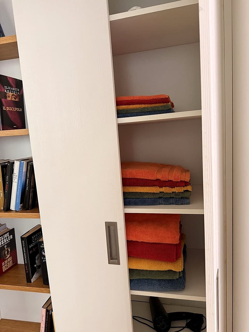 The white cupboard beside the bathroom, open · stacks of towels in bright colours (orange, red, yellow, blue) on three shelves · at the bottom the hairdryer can be glimpsed