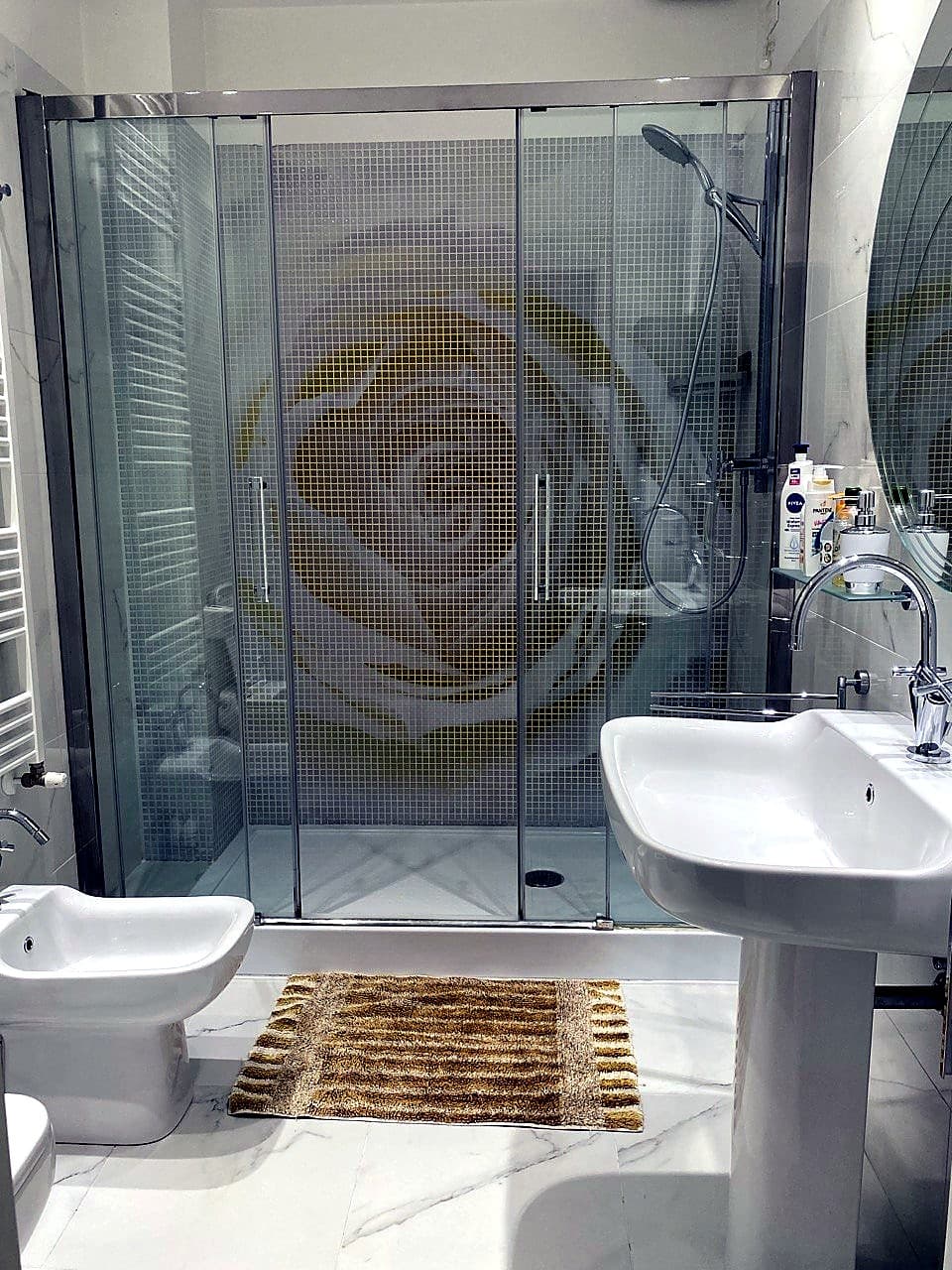 Overall view of the bathroom from the loo area · a handcrafted gold-coloured mosaic shower (a stylised rose effect on the mosaic), a bidet in the foreground, a white wall-hung basin on the right, white marble floor and walls