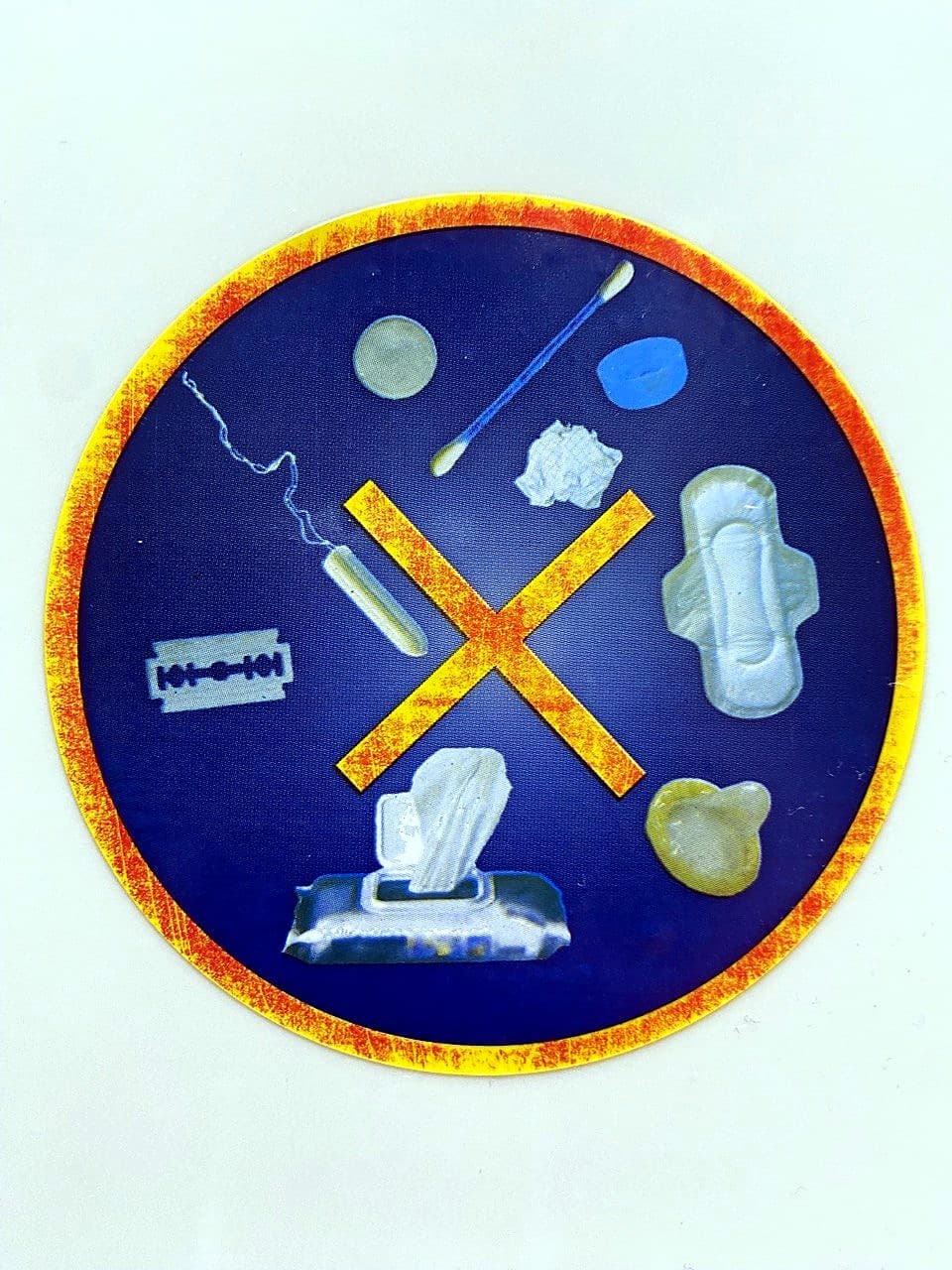 A round sticker above the loo · inside a yellow-orange circle a large golden X crosses out a row of illustrated objects that mustn't be flushed down the loo: cotton buds, wipes, sanitary towel, tampon, nappy, cotton pads, razor