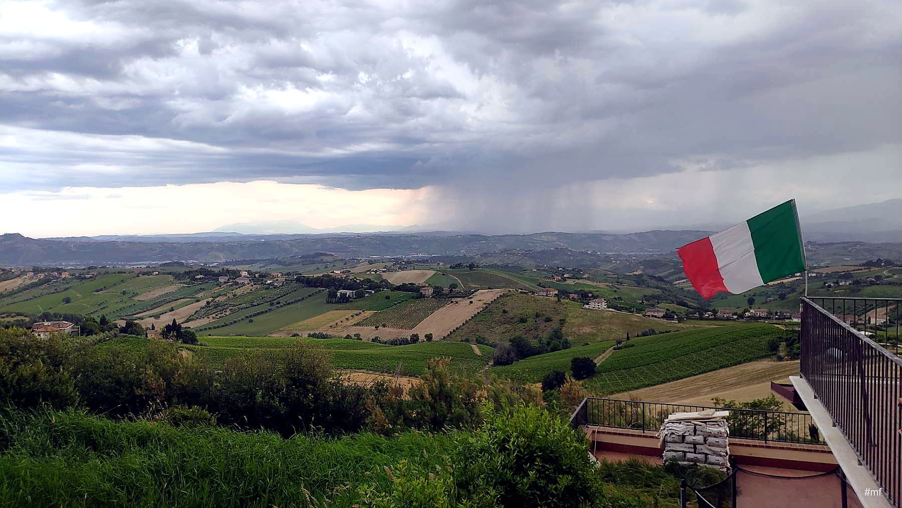 Veduta delle colline picene da Acquaviva Picena · campi coltivati, viti, ulivi, all'orizzonte i Sibillini, bandiera italiana sul belvedere del borgo
