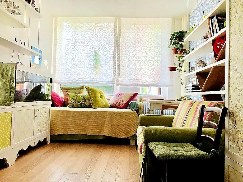 Das zweite Schlafzimmer in La Tana · ein Sofa mit bunten Kissen, links das Aquascape, rechts ein Bücherregal, ein Schreibtisch vor dem Fenster mit einem verzierten Vorhang