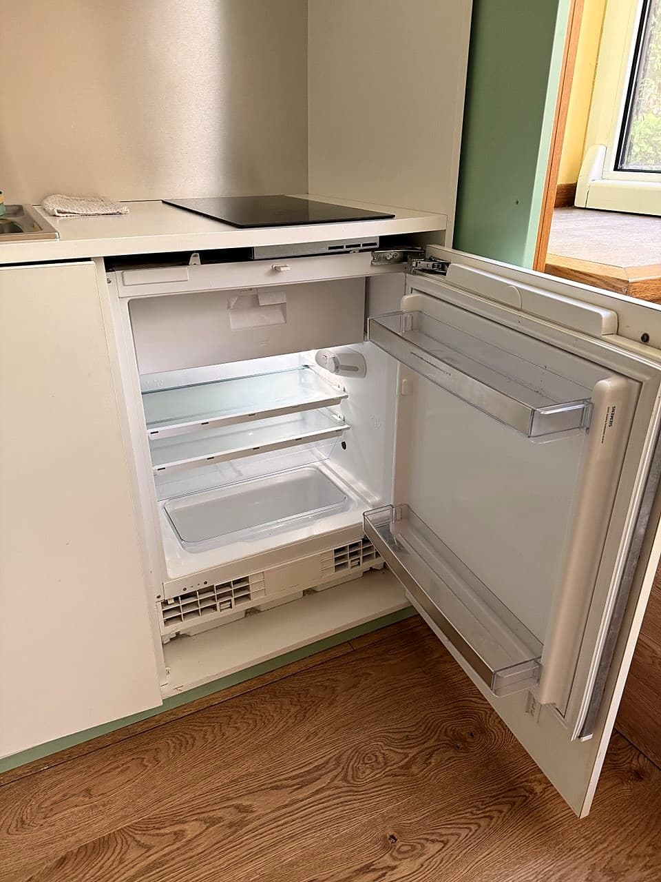Ein kompakter Siemens-Kühlschrank, eingebaut unter dem Induktionskochfeld, geöffnet · zwei Glasablagen und eine Schublade unten