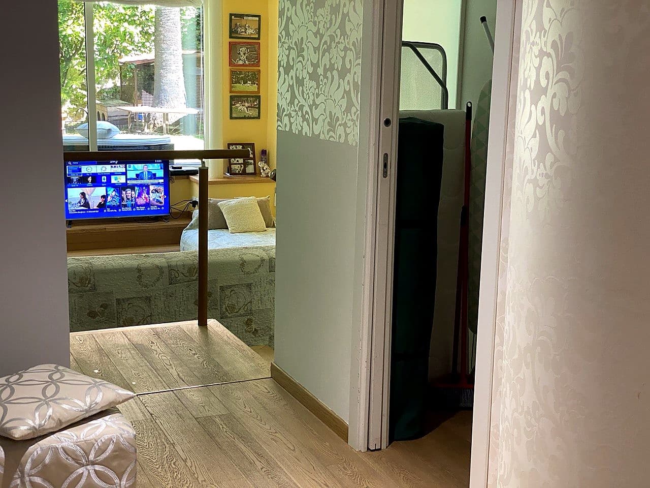 Blick vom Eingang in Richtung Wohnung · der eingeschaltete Smart-TV, ein zurechtgemachtes Schlafsofa unter dem Fenster, und rechts der kleine Vorratsschrank der Waschküche mit der Matratze eines Reisebetts und einem Besen