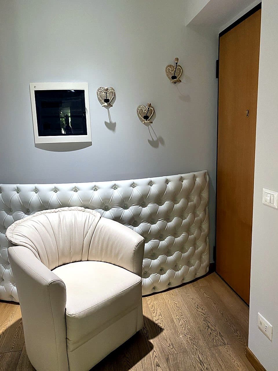 Der kleine Eingangsbereich der Wohnung · ein weißes Sofa mit Knopfheftung, ein abgerundeter Sessel, der Sicherungskasten an der Wand und die helle Holz-Eingangstür rechts