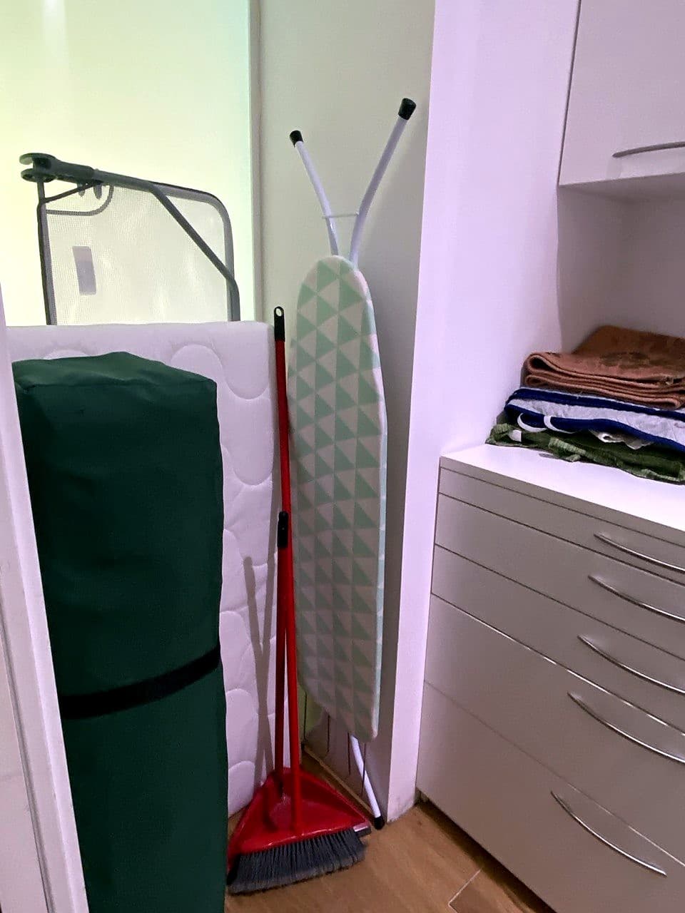 Een hoek van de wasruimte met een strijkplank, een bezem, een opgerolde matras voor het reisbedje en een kolom laden met opgevouwen handdoeken en linnengoed