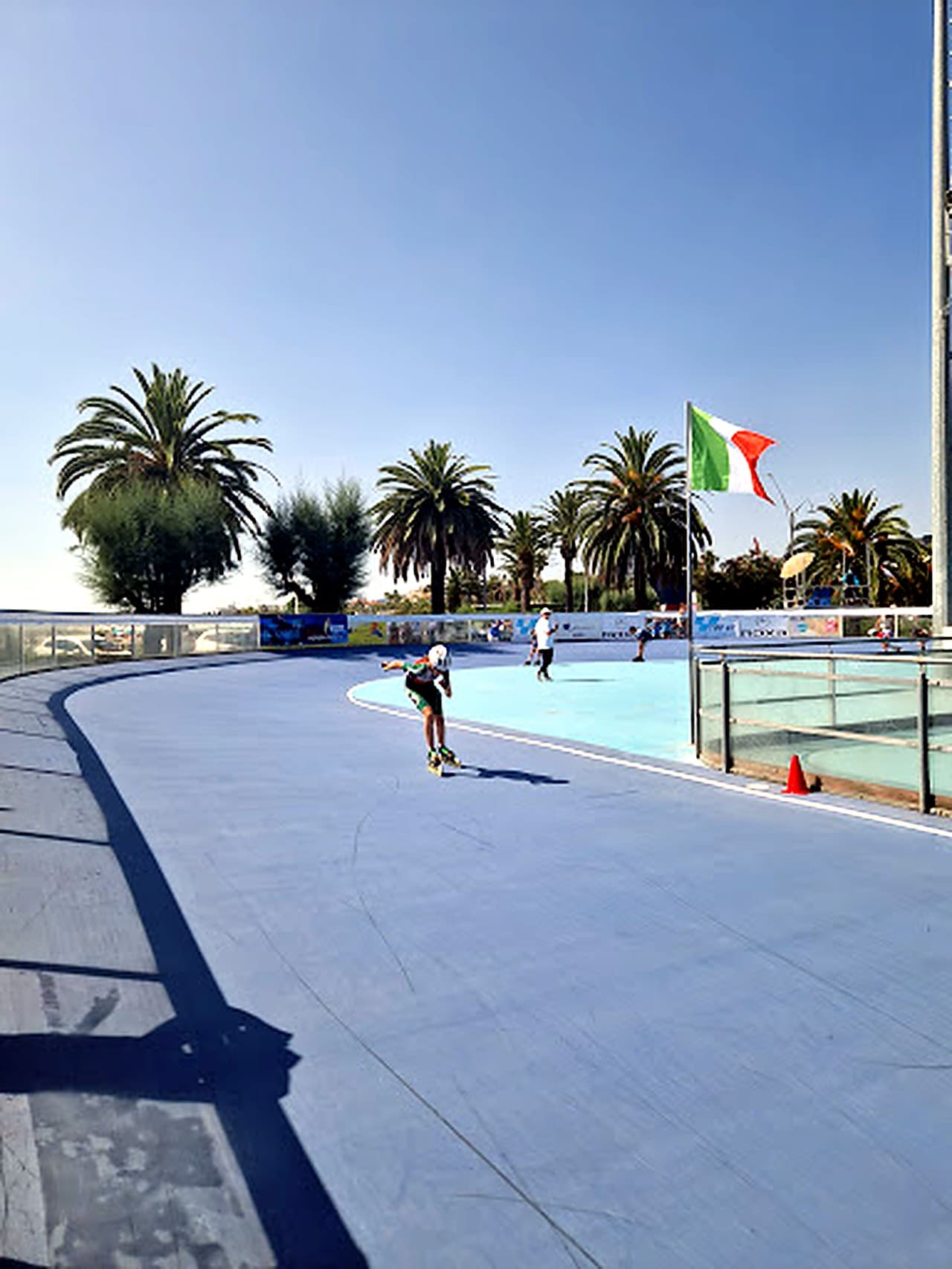 Pista da pattinaggio del Pattinodromo Fernando Panfili a San Benedetto del Tronto · superficie blu, palme e bandiera italiana sullo sfondo, una pattinatrice in allenamento