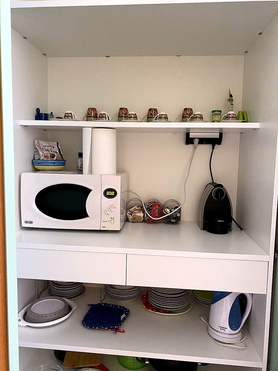 Die geöffnete Tür des Küchenschranks · innen oben die DeLonghi-Mikrowelle und unten der Vorratsschrank mit Küchenrolle, Nudelpackungen, Trinkgläsern, Kaffeekapseln