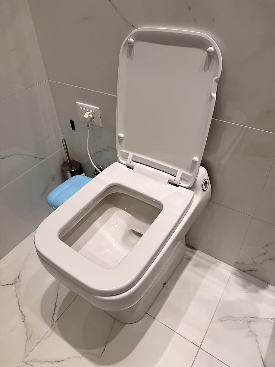 The modern white wall-hung loo, with the lid raised · a power socket visible behind · marble-effect tiled floor